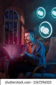 Young Man Using Gadget With Neon Notifications At Home At Night. Sitting On Armchair, Chatting, Talking, Messeging With Friends. Social Media Abusing, Chatting And Browsing Internet, Gadget Addiction.