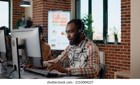 Young Man Using Headphones At Call Center To Help People With Telemarketing Problems. Helpdesk Employee Working At Customer Support Service, Giving Assistance To Business Clients.