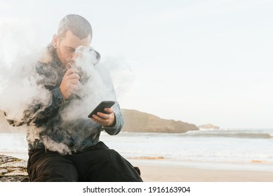 Young Man Using His Electronic Cigarette To Quit Smoking While Consulting His Smart Phone. Person Vaping And Making Clouds Of Vapor.