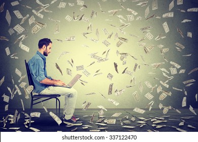 Young man using a laptop building online business making dollar bills cash falling down. Beginner IT entrepreneur under money rain. Success economy concept 