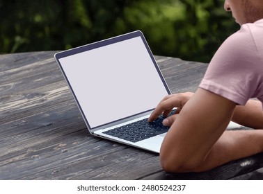 Young man using laptop computer while sitting at wooden table among nature outside. Alternative office place. Blank computer screen, white-screen laptop computer mockup, copy space for text or design - Powered by Shutterstock