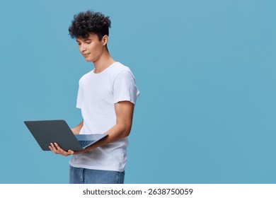 young man using laptop computer wearing casual white t-shirt standing on blue background looking focused and engaged in technology