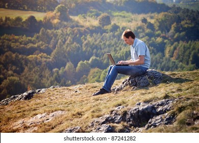 Young Man Using Laptop Sitting On The Grass On The Hillside