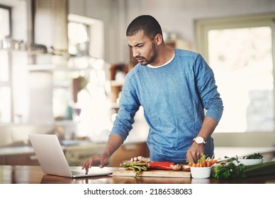 Young Man Using A Laptop While Cooking In A Kitchen, Checking The Internet. Male Watching A Tutorial Online While Preparing A Healthy, Organic Homemade Meal. Guy Following A Easy Vegetarian Recipe