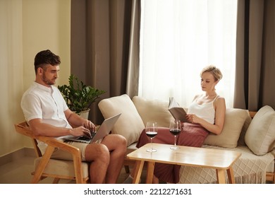 Young Man Using Laptop While Sitting On Armchair With Woman Sitting On Sofa In Front Of Him And Reading A Book