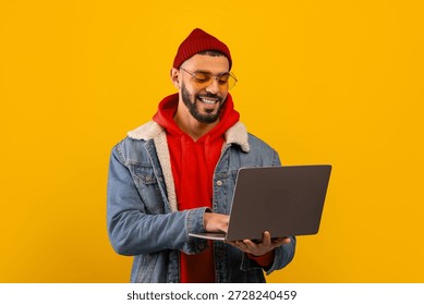 Young Man Using Laptop While Smiling Against a Bright Yellow Background in a Casual Outfit