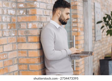 Young man using a laptop while standing, leaning against a brick wall, engaged in remote work or online business