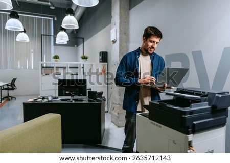 Young man using laptop while standing next to printer in modern office setting during daytime