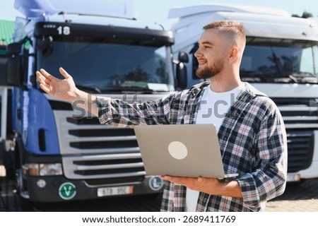 Young man using laptop while managing logistics for truck fleet, standing outdoors beside semitrailers