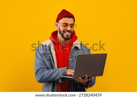 Young Man Using Laptop While Smiling Against a Bright Yellow Background in a Casual Outfit