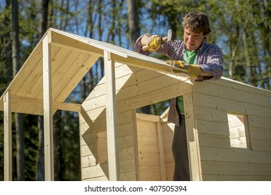 Young Man Using A Mallet To Fix A Nail Into A Roof Of A Wooden Playhouse For Children In A DIY Concept.