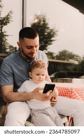 Young man using a phone mobile for work and holding his son on his hand on the cafe. Being a daddy and working everywhere. Freelancing and networking concept. - Shutterstock ID 2364785097