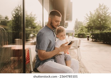 Young man using a phone mobile for work and holding his son on his hand on the cafe. Being a daddy and working everywhere. Freelancing and networking concept. - Shutterstock ID 2364785101
