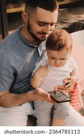 Young man using a phone mobile for work and holding his son on his hand on the cafe. Being a daddy and working everywhere. Freelancing and networking concept. - Shutterstock ID 2364785113