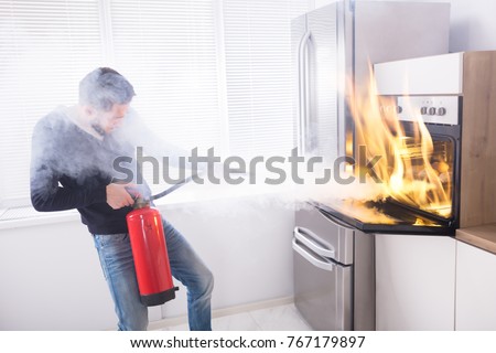 Young Man Using Red Fire Extinguisher To Stop Fire Coming From Oven In Kitchen