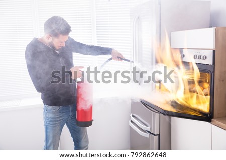 Young Man Using Red Fire Extinguisher To Stop Fire Coming From Oven In Kitchen