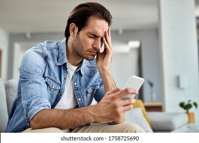 Young man using smart phone and holding his head in pain at home.