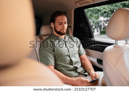 Young man using smartphone in the back seat of a car, wearing earbuds, relaxed atmosphere with soft lighting