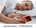 Young man using smartphone in bed, checking social media, experiencing digital overload