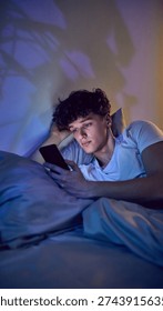 Young man using smartphone in bed at night with blue screen light. Concept of late night social media use, mobile apps engagement, digital consumption and screen time habits.
