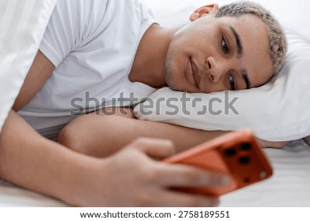 Young man using smartphone in bed, checking social media, experiencing digital overload