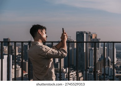 Young man using smartphone to capture the expansive cityscape from a high-rise building balcony in Bangkok, Thailand, enjoying travel and technology