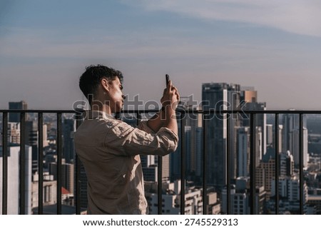 Young man using smartphone to capture the expansive cityscape from a high-rise building balcony in Bangkok, Thailand, enjoying travel and technology