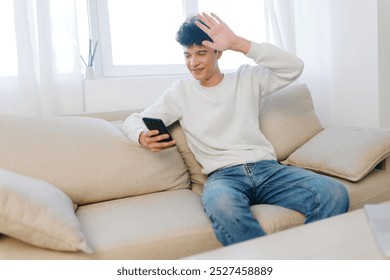 young man using smartphone, casual lifestyle, comfortable home setting, beige sofa, natural light, modern interior, relaxed atmosphere, friend greeting through video call, smiling expression - Powered by Shutterstock