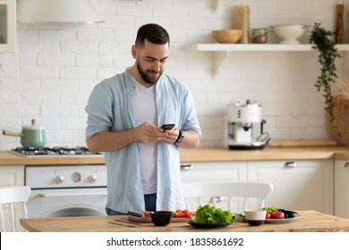 Young man using smartphone, cooking salad in modern kitchen, searching vegetarian snack recipe in internet, looking at phone screen, interested male preparing dinner, fresh vegetables