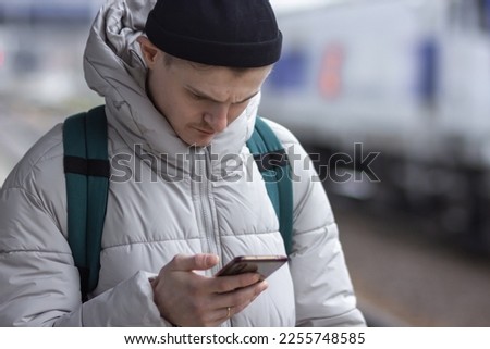 Young man using smartphone, texting, cheks the route of trip on google mapes while waiting train at railway station.