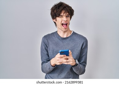 Young Man Using Smartphone Typing A Message Winking Looking At The Camera With Sexy Expression, Cheerful And Happy Face. 