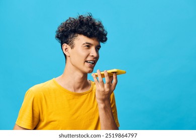 young man using smartphone for voice commands, bright blue background, cheerful expression, engaging in conversation, modern communication concept, technology and youth interaction - Powered by Shutterstock