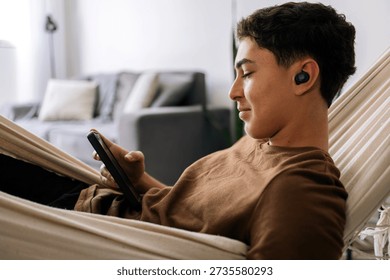 Young man using a smartphone while lying in a hammock at home with wireless earbuds. Concepts of leisure, technology, comfort and modern lifestyle.
