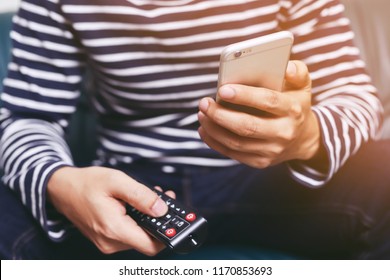 Young Man Using Television Remote Control And Mobile Smart Phone  Is Sitting On A Sofa. On Or Off Tv. 