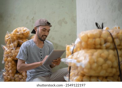 A young man is utilizing a tablet in a storage area that is filled with numerous bags of snacks