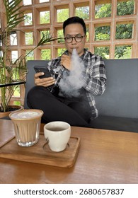 Young man vaping while using smartphone at coffee shop with latte and espresso on wooden table, relaxing lifestyle concept