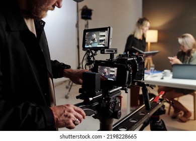 Young man with videocamera standing in studio in front of his assistant with documents and female model taking part in commercial shooting