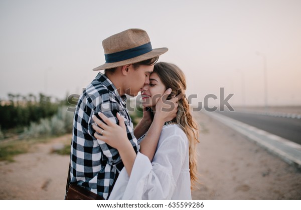 彼女の顔を優しく持ち 夜明けに道端に立って彼女にキスをするビンテージハットをかぶった若い男性 愛するカワイイ夫婦が外で抱き合う美しいカップルは ロマンチックな夜のデートを楽しむ の写真素材 今すぐ編集