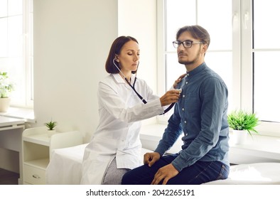 Young man visiting doctor for health exam. Serious middle aged female physician with stethoscope sitting on modern examination couch and listening to male patient's lungs or checking his heartbeat