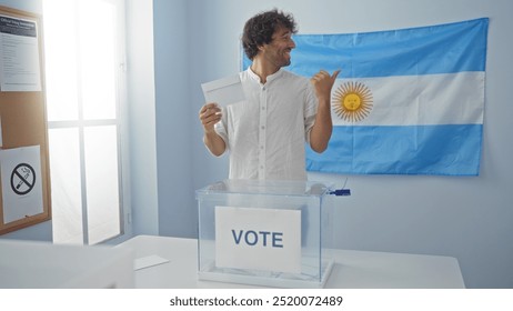 Young man voting in an electoral room with an argentinian flag in the background, holding a ballot and pointing while smiling - Powered by Shutterstock