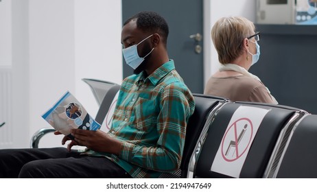 Young Man Waiting In Hospital Reception Lobby To Start Consultation With Medic During Coronavirus Pandemic. Diverse People Sitting In Waiting Area Chairs To Do Covid 19 Consultation.