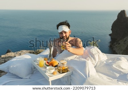 A young man wakes up alone in a white bed on a cliff overlooking the sea