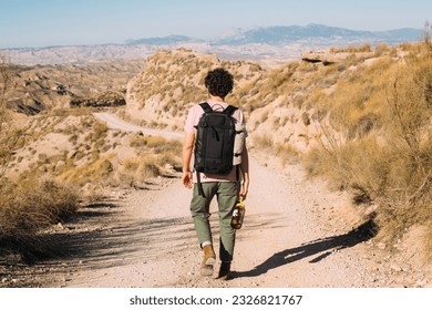 Young man walk away from camera, in leather boots, wear black backpack and carry water flask on empty dusty desert road. Moody solo adventure. Hot sunny day for a hike - Powered by Shutterstock