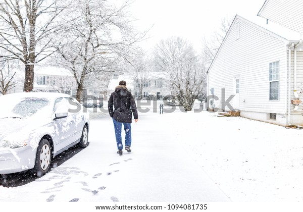 Young man walking by car in driveway in\
neighborhood with snow covered ground during blizzard white storm,\
snowflakes falling in Virginia suburbs, single family homes to\
check mail in mailbox