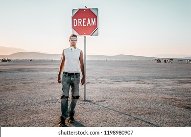 Young Man Walking By The Dream Sign In The Middle Of A Desert At The Burning Man - Art And Music Festival. Sign To Keep Dreaming.