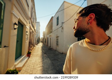 A Young Man Is Walking Down A Narrow Mediterranean Street Looking Over His Shoulders Into The Sunset Breaking Through The Houses Behind Him.