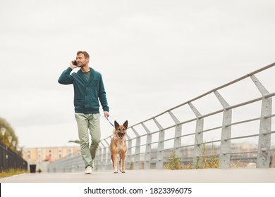 Young Man Walking With His Dog Along The Street And Talking On Mobile Phone