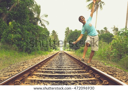 Young man walking on railroad track while listening to music with headphones on his smartphone in rural area in Bentota, Sri Lanka. Vintage effect. Risk, reckless, danger, dare, no fear concepts