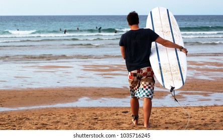 Young Man Walking Towards The Ocean Carrying A Surfboard And Wearing Cool Boardshorts