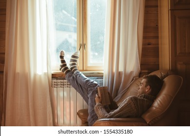 Young Man In Warm Sweater Reading Book While Relaxing On Armchair By The Window And Electric Radiator Inside Cozy Log Cabin